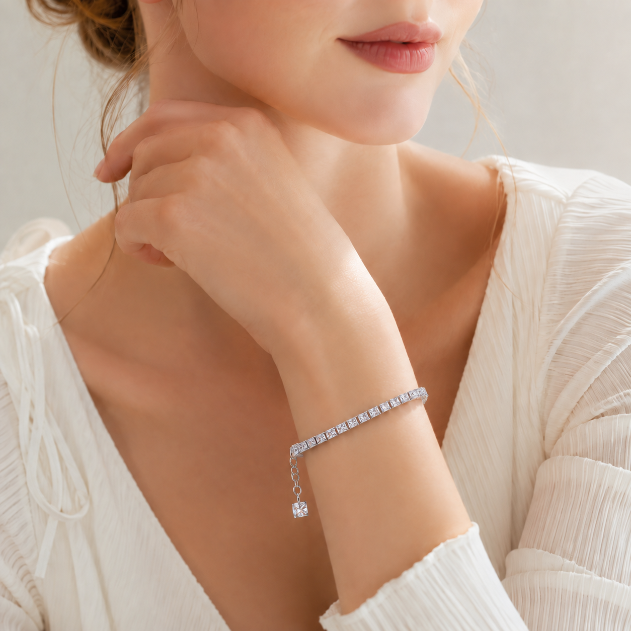 A close-up of a woman wearing a delicate silver bracelet with a row of clear square stones and a small dangling charm, paired with a white ribbed top.