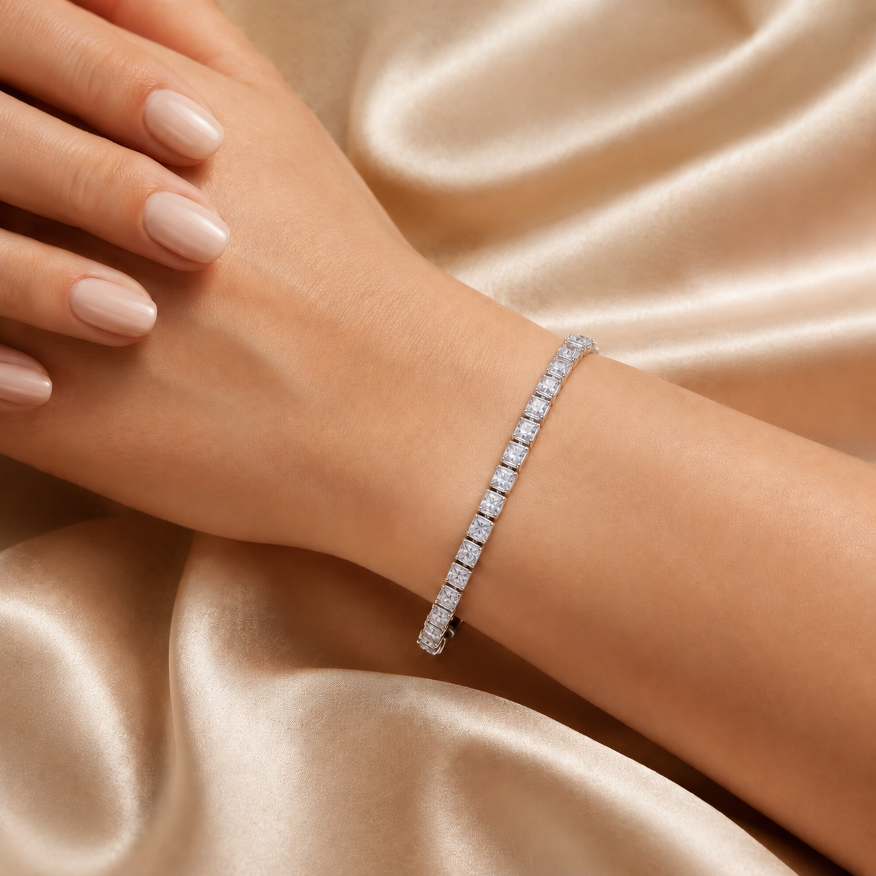 A close-up of a woman's wrist and hand wearing a slim diamond tennis bracelet, posed against a draped champagne satin fabric background.